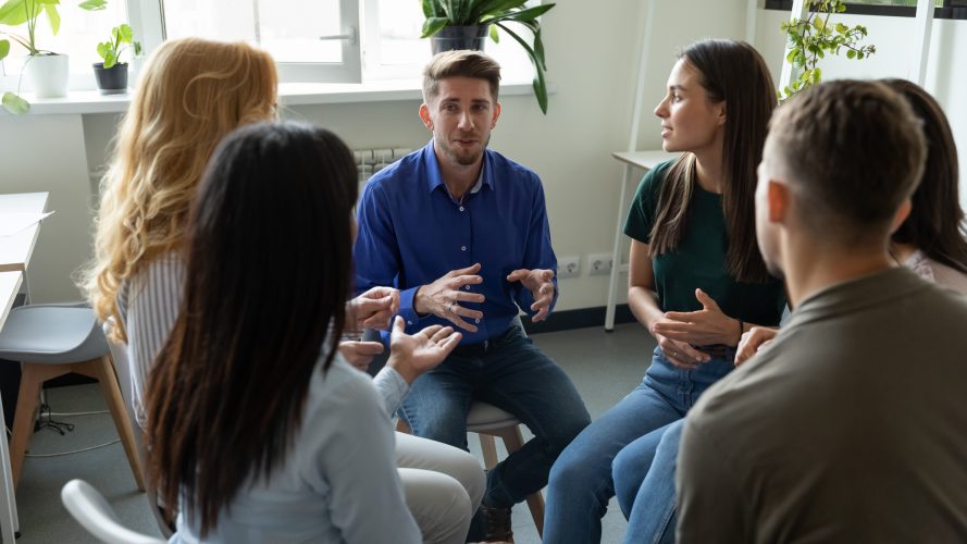 Confident male coach leader talking holding briefing for team employees in office, explaining business strategy, plan to subordinates. Businessman sitting at company meeting with diverse colleagues.